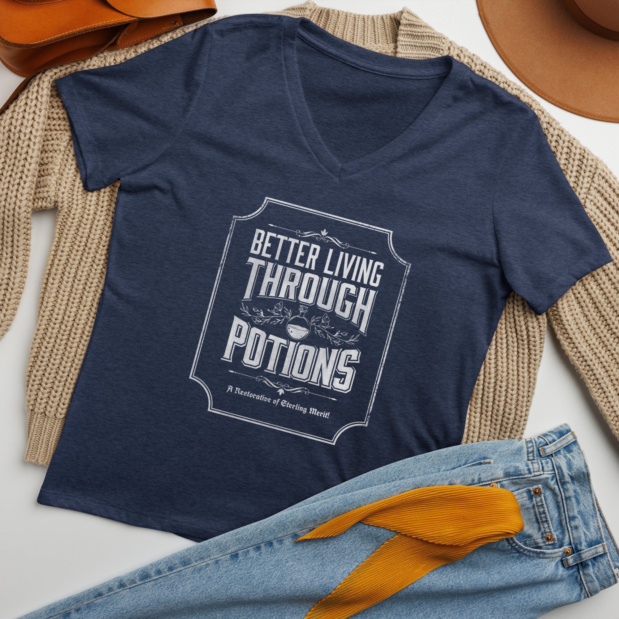 A navy blue V-neck t-shirt with a white graphic print reading 'Better Living Through Potions' laid flat on a beige knit sweater, with blue jeans and a mustard yellow ribbon in the foreground.
