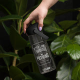 A hand with lavender nail polish holds a CamelBak water bottle against a dark background with green foliage. The bottle is clear  grey with a black lid and strap, featuring a black and white label that reads 'Better Living Through Potions' and 'CamelBak'.