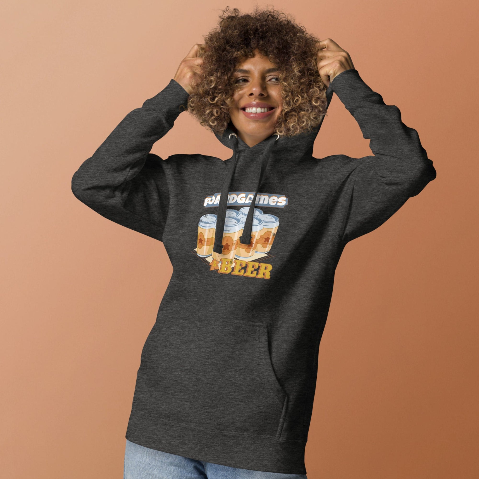A woman with curly hair wearing a dark gray heather hoodie with a graphic print of six beer cans and the text Boardgames and Beer. She is posing with her hands on her head against a solid peach-colored background.