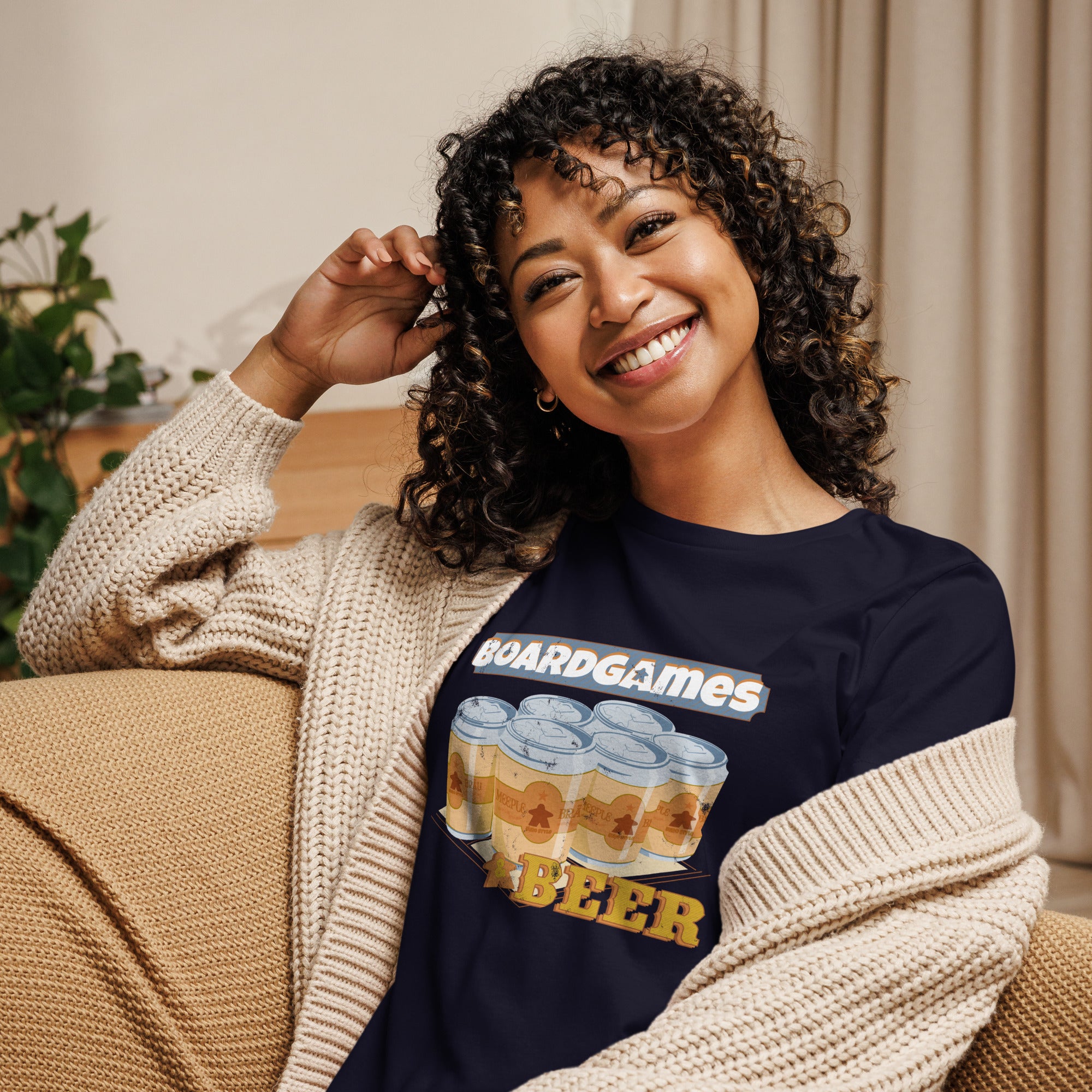 A smiling woman with curly hair sits on a beige couch wearing a navy graphic T-shirt with 'BOARDGAMES BEER' and beer cans, layered under a cream knit cardigan.