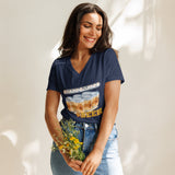 A woman wearing a navy blue v-neck t-shirt with a 'Board Games' graphic featuring beer cans, paired with light-wash blue jeans, holding a small bouquet of yellow flowers against a sunlit cream wall.