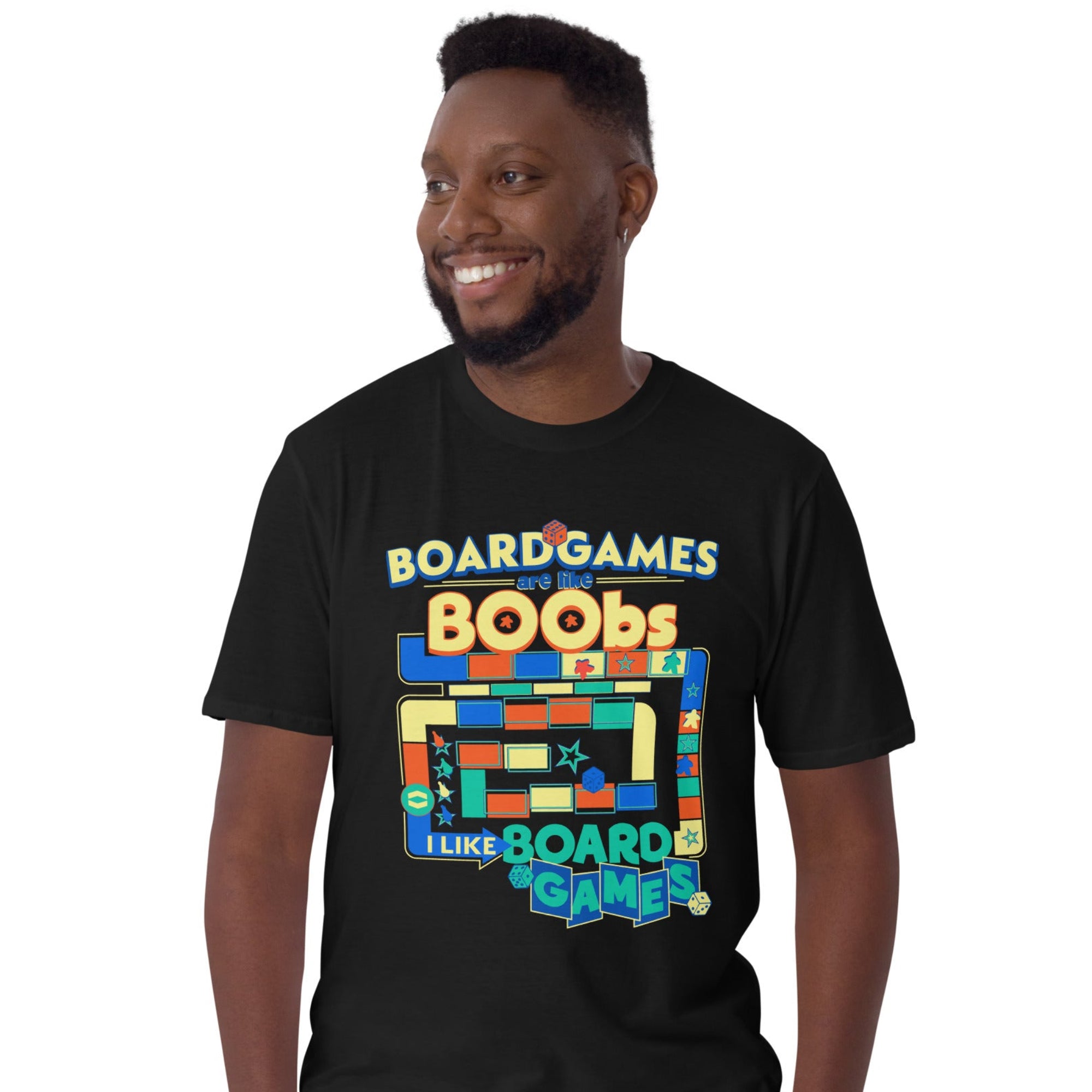 A man wearing a black graphic t-shirt with a colorful retro board game design that reads 'BOARD GAMES ARE LIKE BOOBS I LIKE BOARD GAMES', paired with khaki pants, against a plain white background.