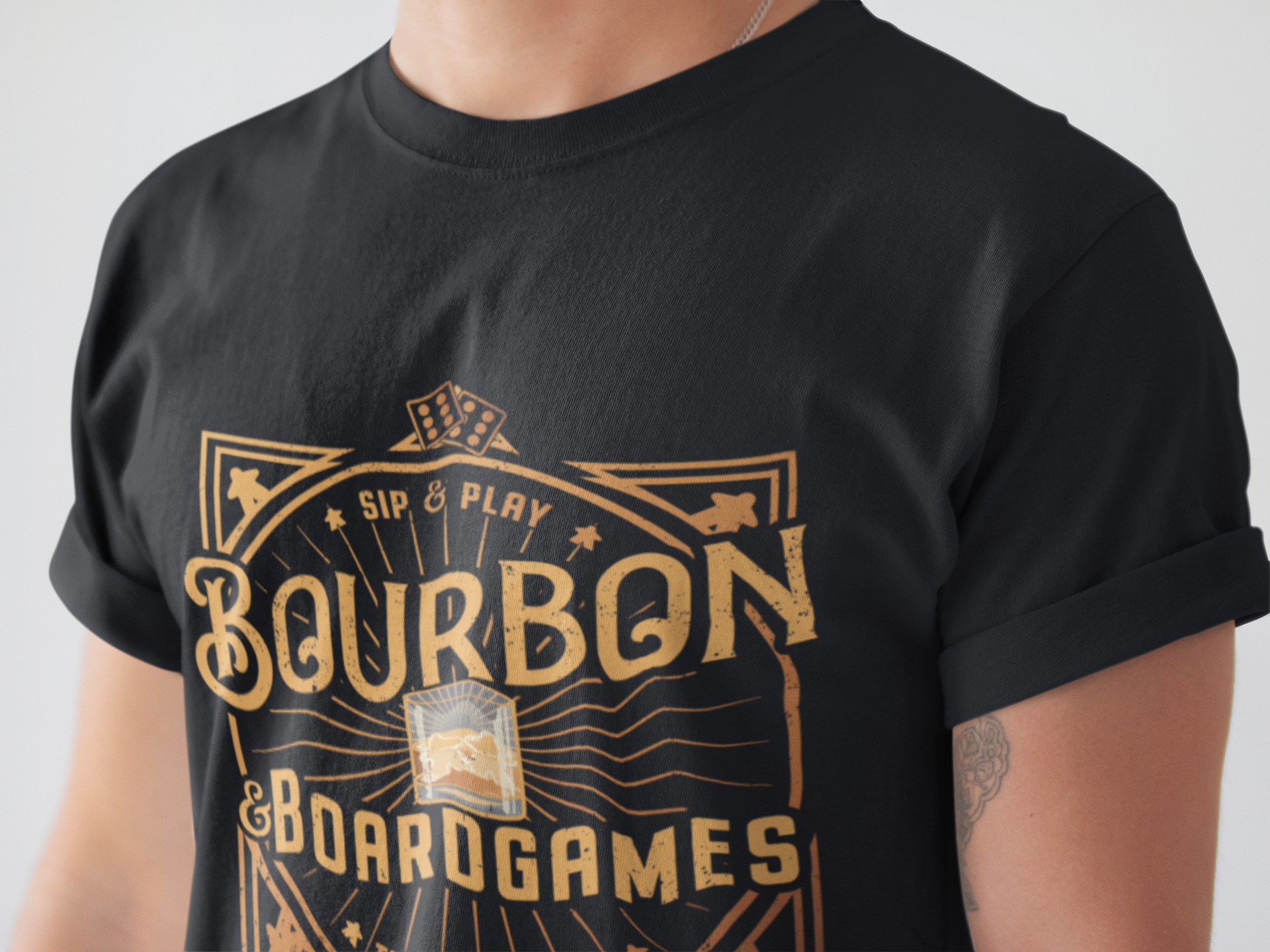 A close-up of a person wearing a black short-sleeve T-shirt with a large gold graphic on the front that reads 'Bourbon & Boardgames' and 'Sip & Play'.