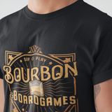 A close-up of a person wearing a black short-sleeve T-shirt with a large gold graphic on the front that reads 'Bourbon & Boardgames' and 'Sip & Play'.