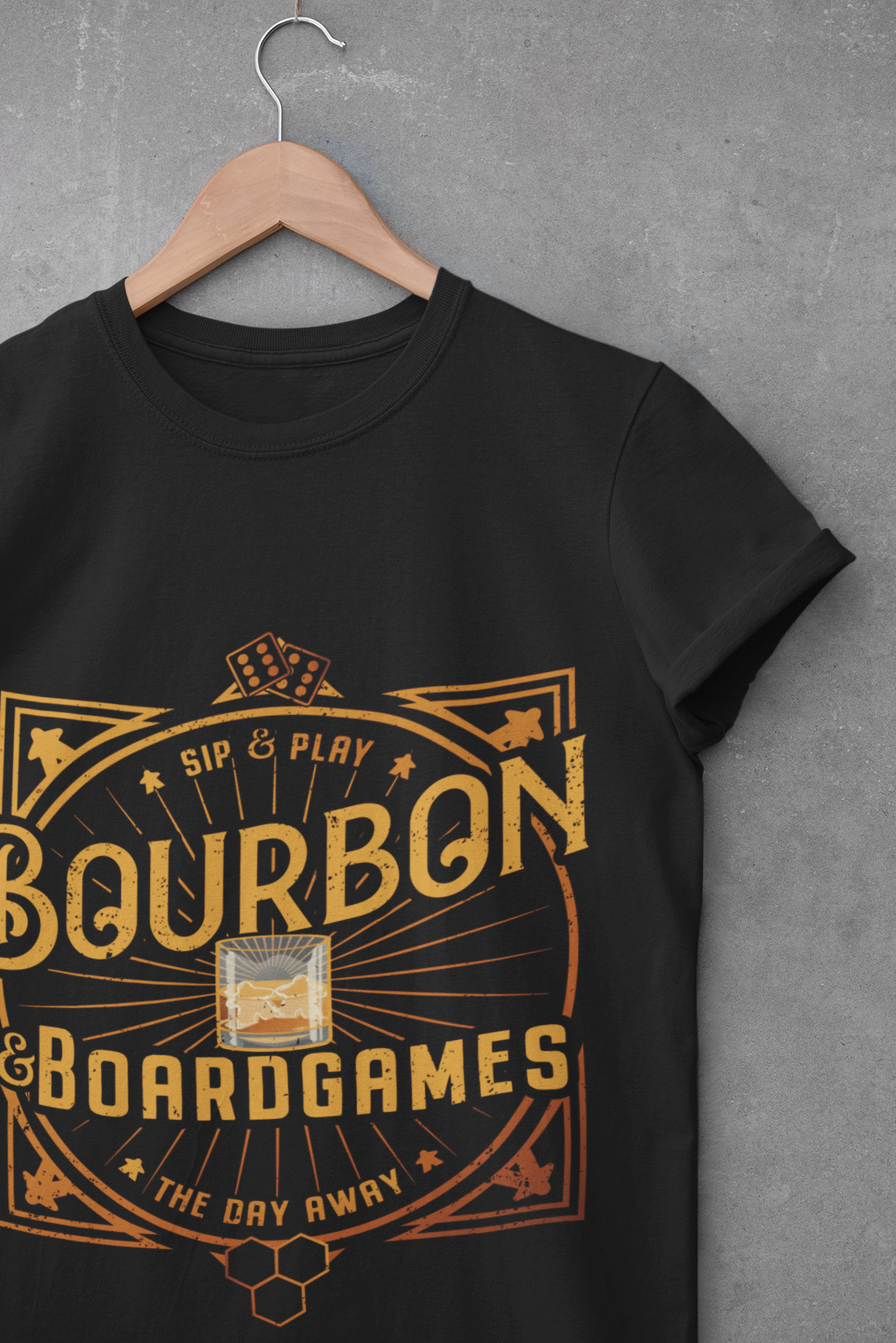 A black short-sleeve t-shirt on a wooden hanger against a textured gray wall. The shirt features a large vintage-style graphic in gold and orange reading 'Bourbon & Boardgames' with 'Sip & Play' and 'The Day Away' around a whiskey glass illustration.