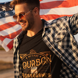 A man wearing round sunglasses, a black graphic t-shirt, and an open black-and-white plaid flannel shirt, standing outdoors with an American flag waving in the background.