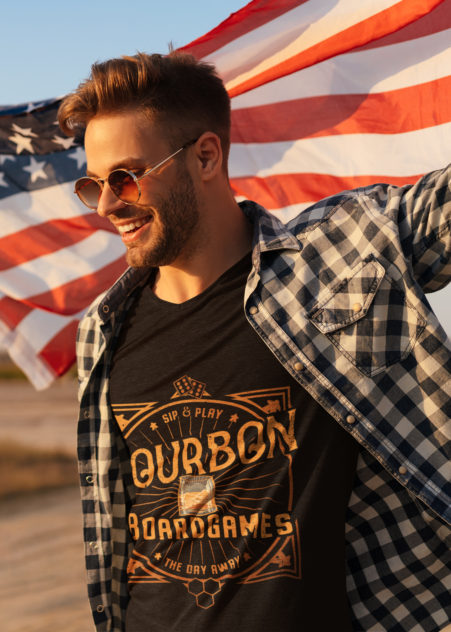 A man wearing round sunglasses, a black graphic t-shirt, and an open black-and-white plaid flannel shirt, standing outdoors with an American flag waving in the background.