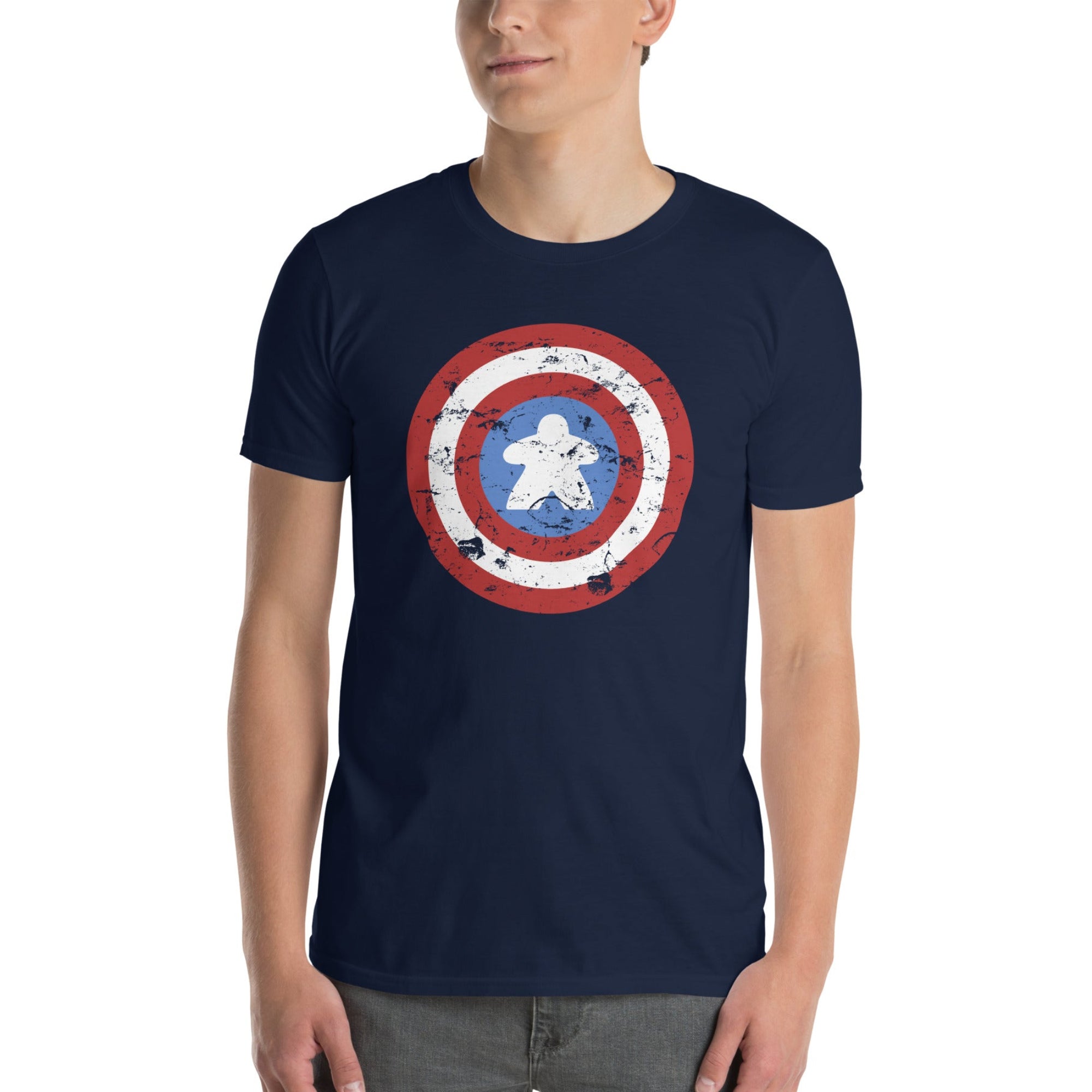 A man wearing a navy blue crewneck t-shirt with a distressed shield graphic on the chest with a meeple at the center, paired with gray jeans, against a plain white background.