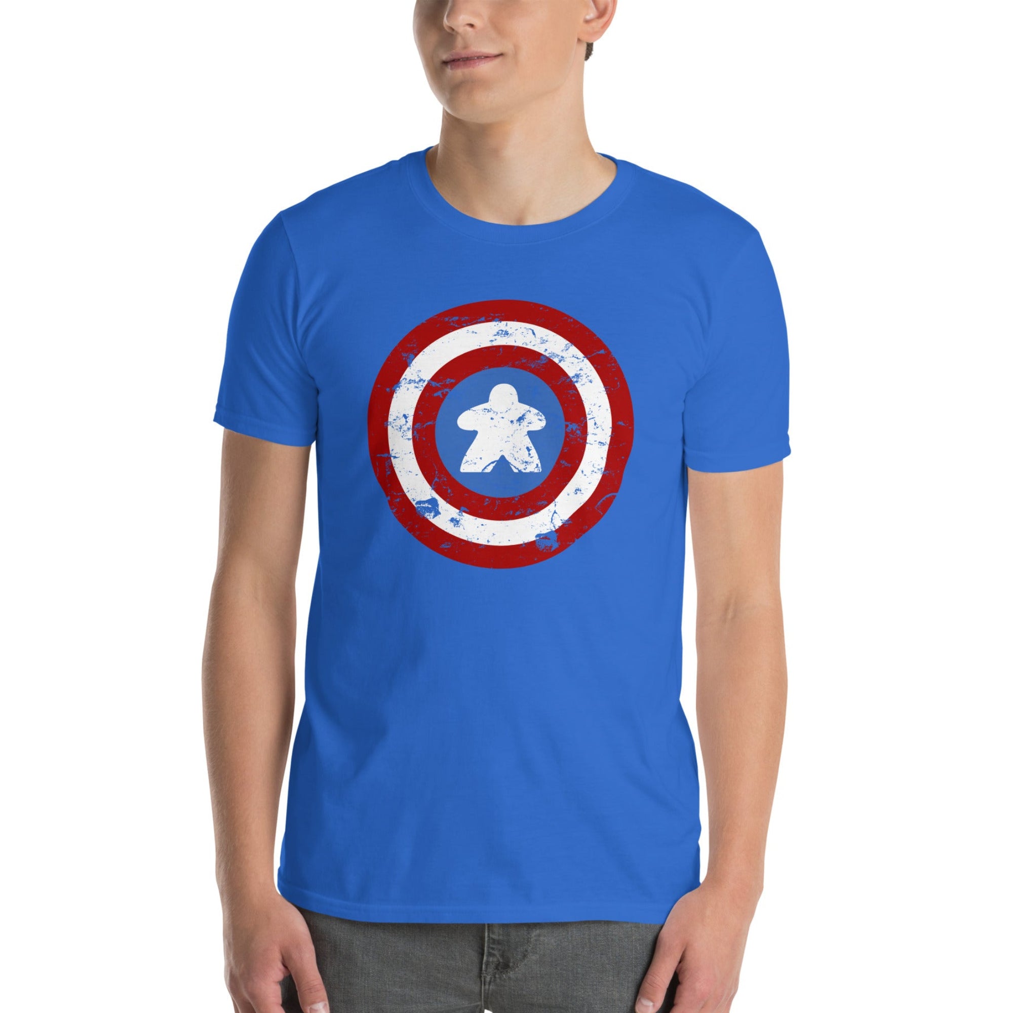 A man wearing a royal blue crewneck t-shirt with a distressed shield graphic on the chest with a meeple at the center, paired with gray jeans, against a plain white background.