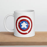 A ceramic coffee mug printed with a red, white, and blue shield design featuring a meeple at its center in place of a star.