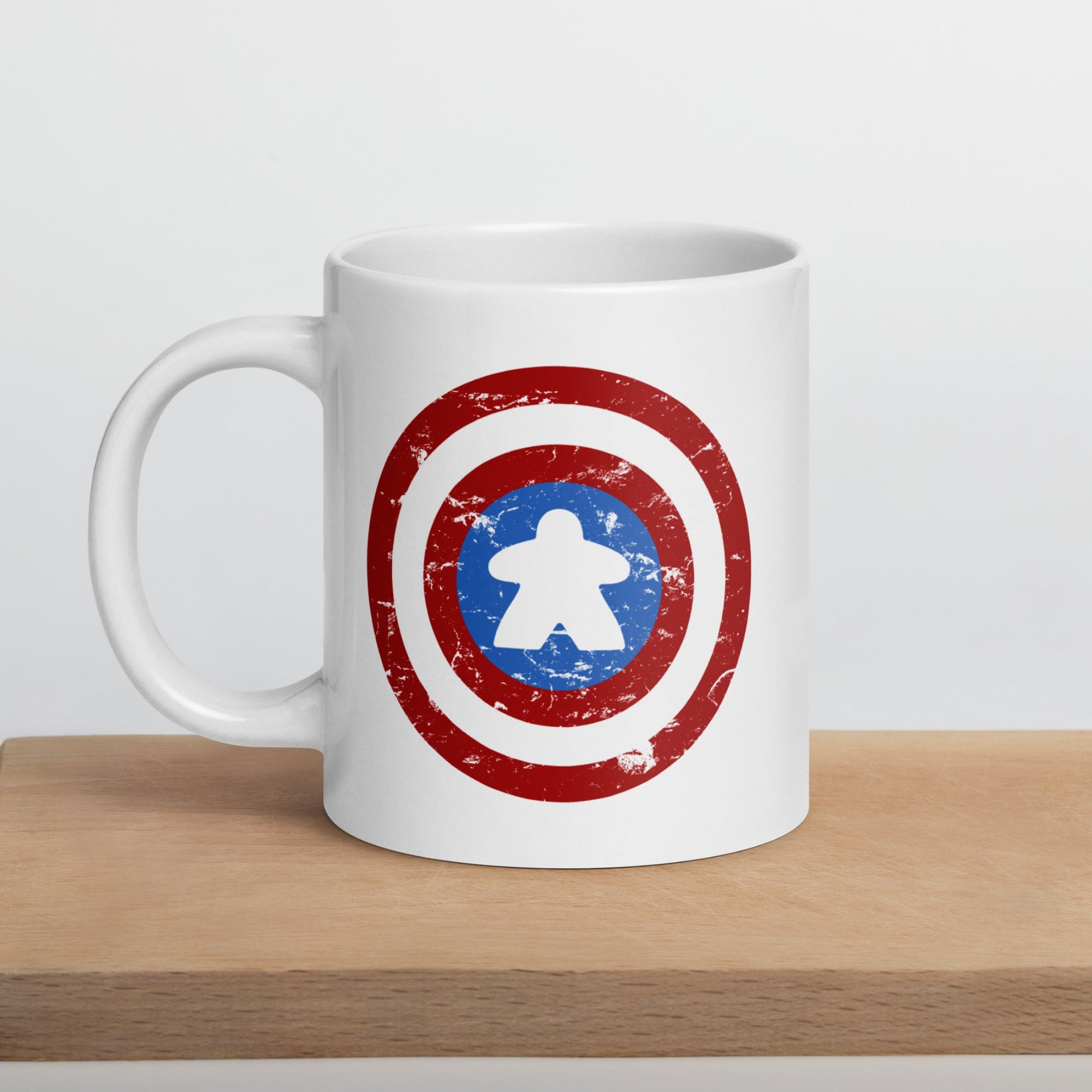 A ceramic coffee mug printed with a red, white, and blue shield design featuring a meeple at its center in place of a star.