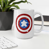 A white ceramic mug with a distressed Captain Meeple shield graphic, displayed on a white desk next to a keyboard, a potted plant, and a book.