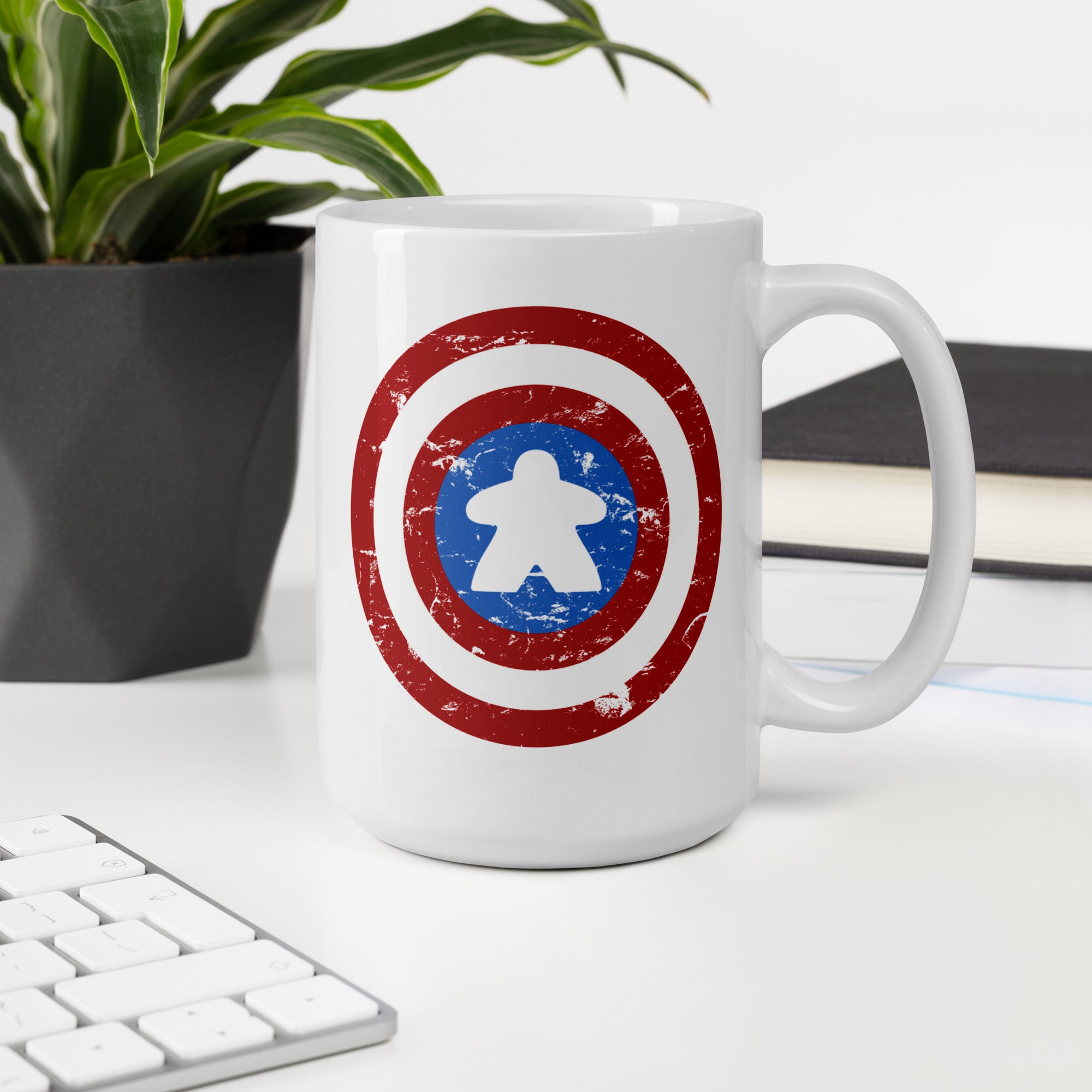A white ceramic mug with a distressed Captain Meeple shield graphic, displayed on a white desk next to a keyboard, a potted plant, and a book.