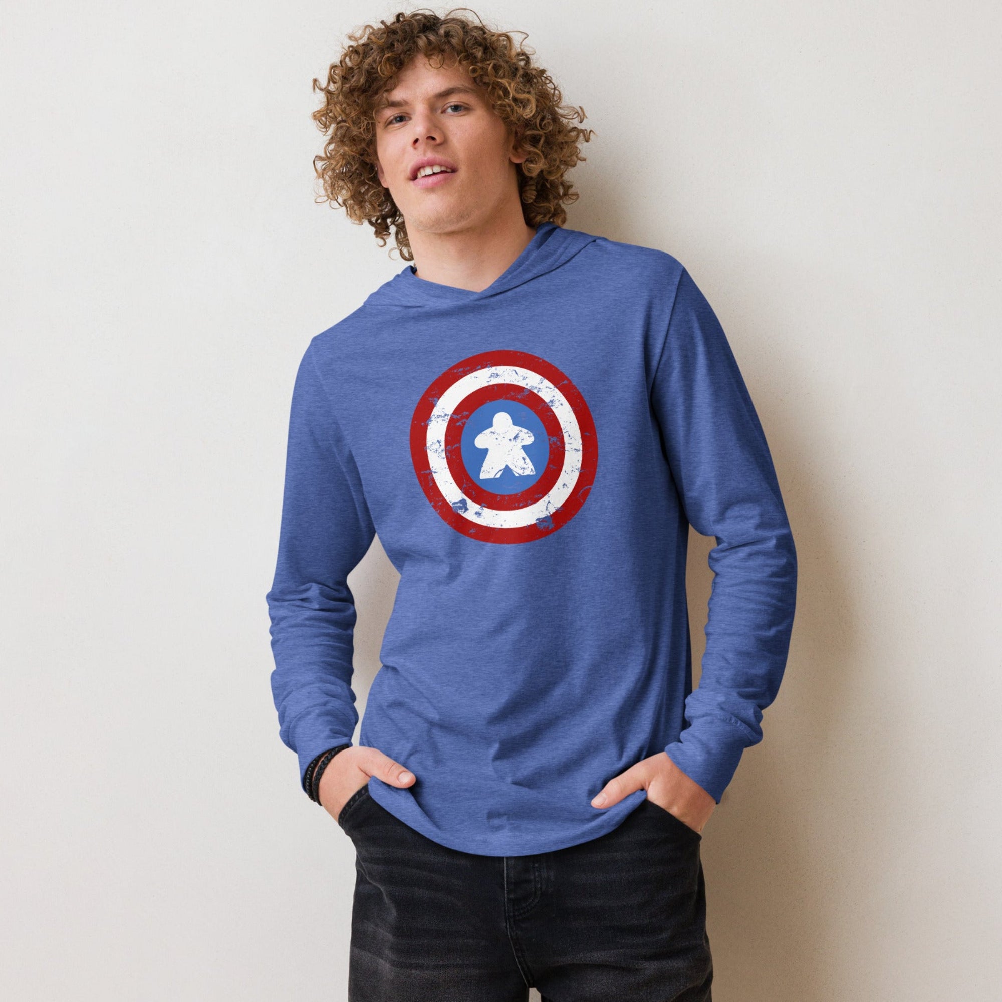 A young man with curly brown hair wearing a blue long-sleeve hooded shirt with a captain meeple shield on the chest, paired with black denim shorts, standing against a plain off-white wall.