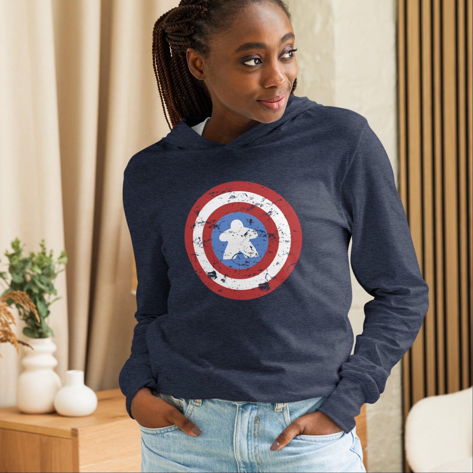 A woman wearing a navy blue hoodie with a distressed Captain Meeple shield graphic on the front, paired with light blue jeans, standing in a cozy indoor setting with beige curtains and wooden paneling.