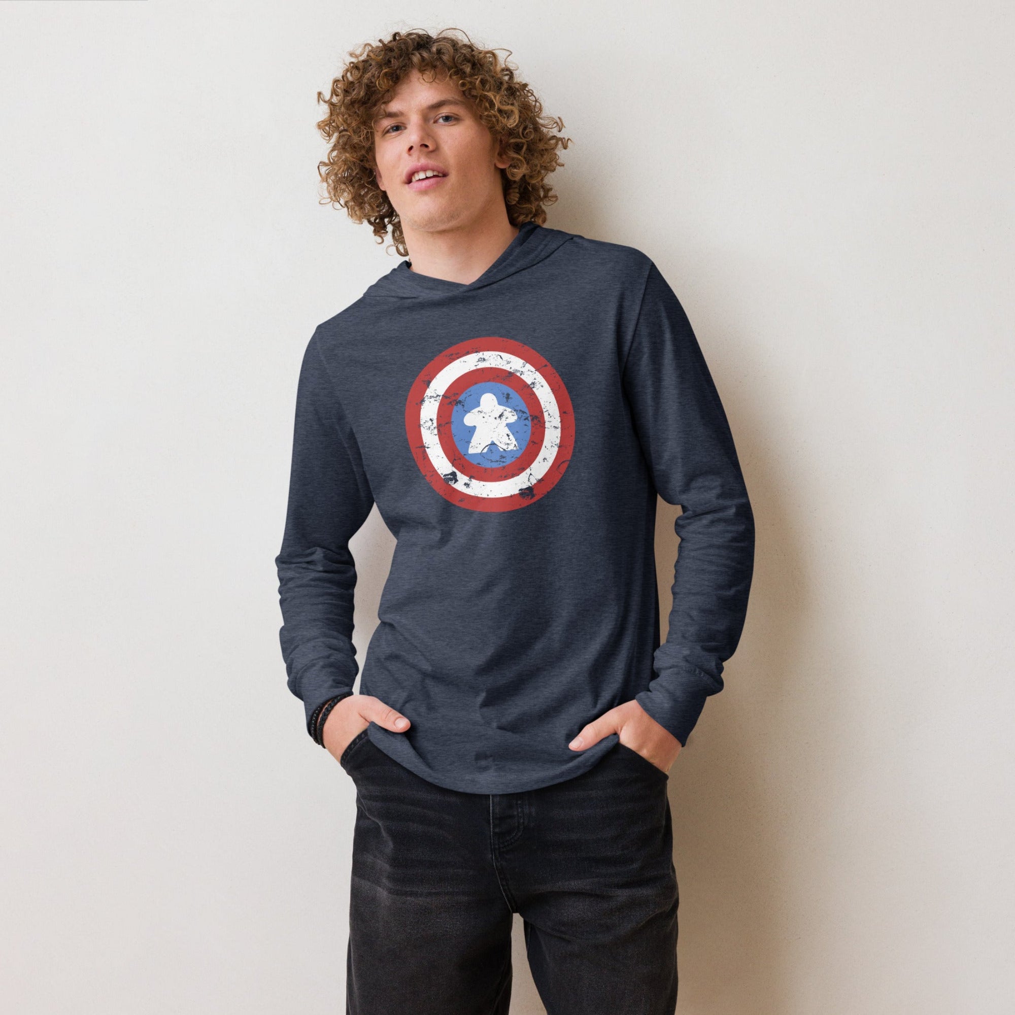 A young man with curly light brown hair wearing a navy blue long-sleeve hoodie with a Captain Meeple shield graphic on the chest, paired with black denim shorts, standing against a plain off-white wall.
