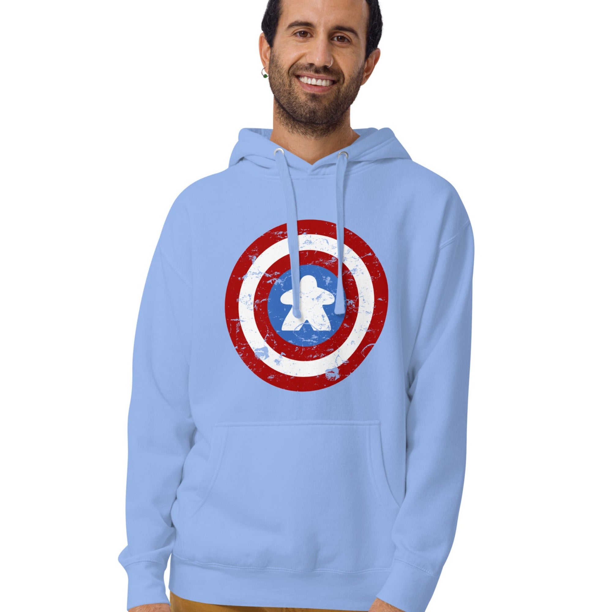 A man wearing a light blue pullover hoodie with a distressed Captain Meeple shield graphic on the chest, paired with brown pants, against a plain white background.