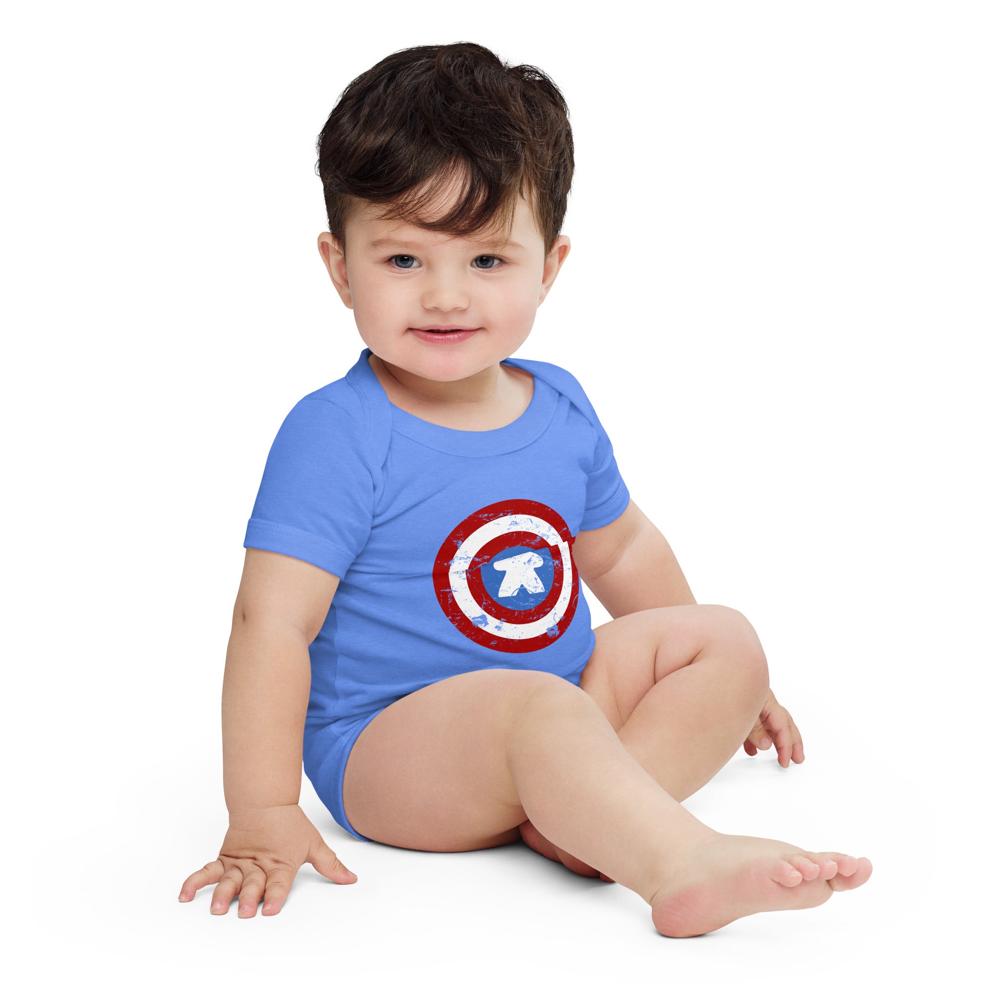 A baby sitting on a white studio background wearing a blue short-sleeve bodysuit with a Captain Meeple shield graphic on the chest.