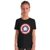A young girl with braided hair stands against a plain white background wearing a black short-sleeve t-shirt with a distressed Captain Meeple shield graphic on the chest.
