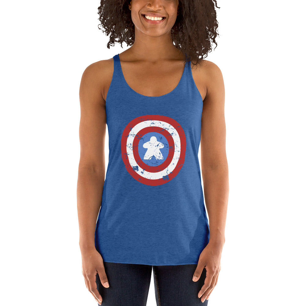 A woman wearing a heather blue racerback tank top with a distressed Captain Meeple shield graphic on the front featuring a white meeple at the center instead of a star, paired with dark jeans, against a plain white background.