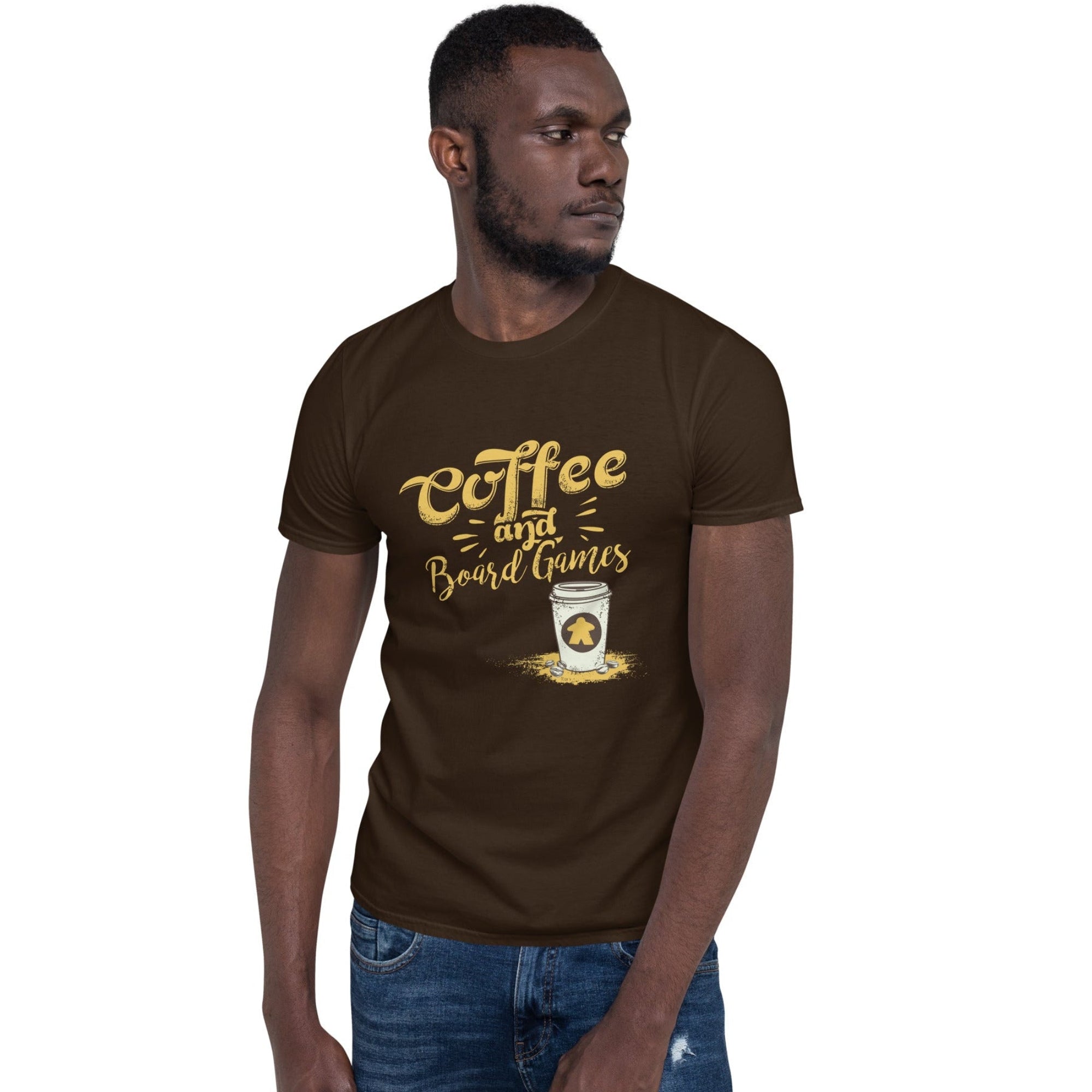 A man wearing a dark brown t-shirt with a graphic that reads 'Coffee and Board Games' in gold lettering, paired with blue jeans, set against a plain white background. There is a white travel mug with a yellow meeple on it.