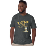 A man wearing a dark heather gray t-shirt with a gold graphic that reads 'Coffee and Board Games' next to a coffee cup with a meeple on it, paired with khaki pants, set against a plain white background.