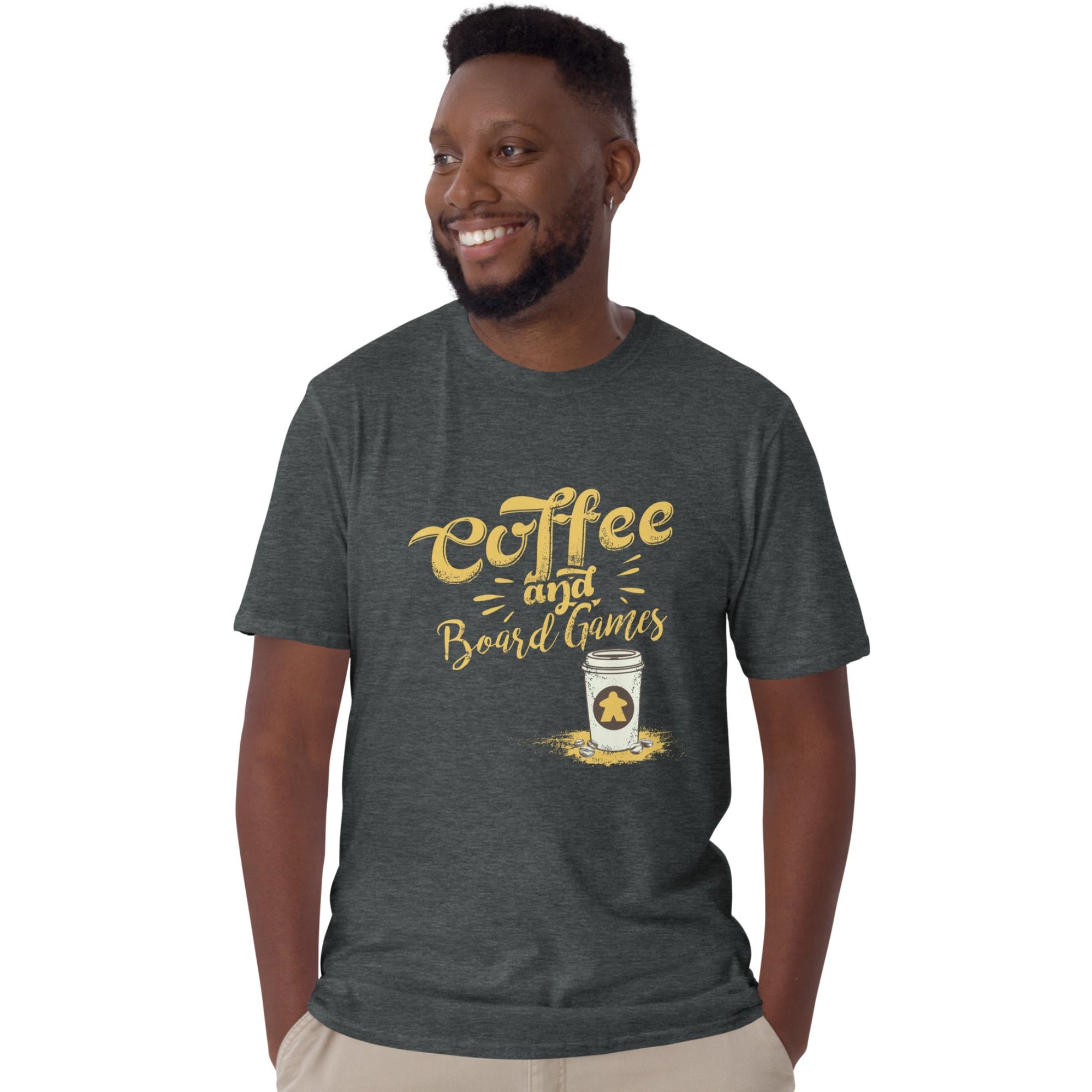 A man wearing a dark heather gray t-shirt with a gold graphic that reads 'Coffee and Board Games' next to a coffee cup with a meeple on it, paired with khaki pants, set against a plain white background.